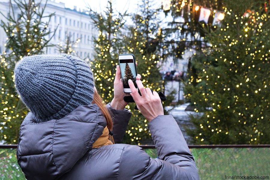 Frau macht mit den Smartphone ein Foto von einem mit Lichtern geschmückten Weihnachtsbaum