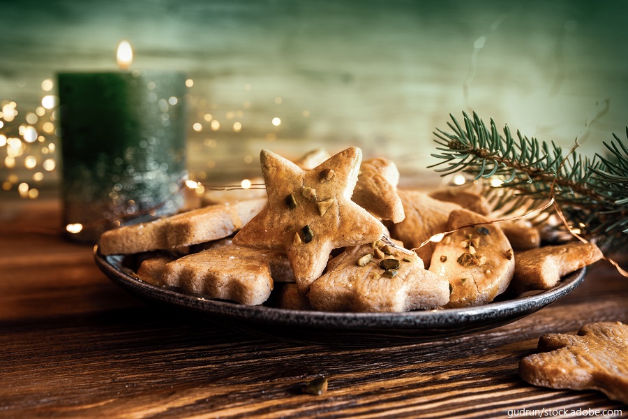 Ein Teller mit Weihnachtsplätzchen in Sternform, umgeben von einer grünen Kerze und Tannenzweigen, dekoriert mit Lichtern.