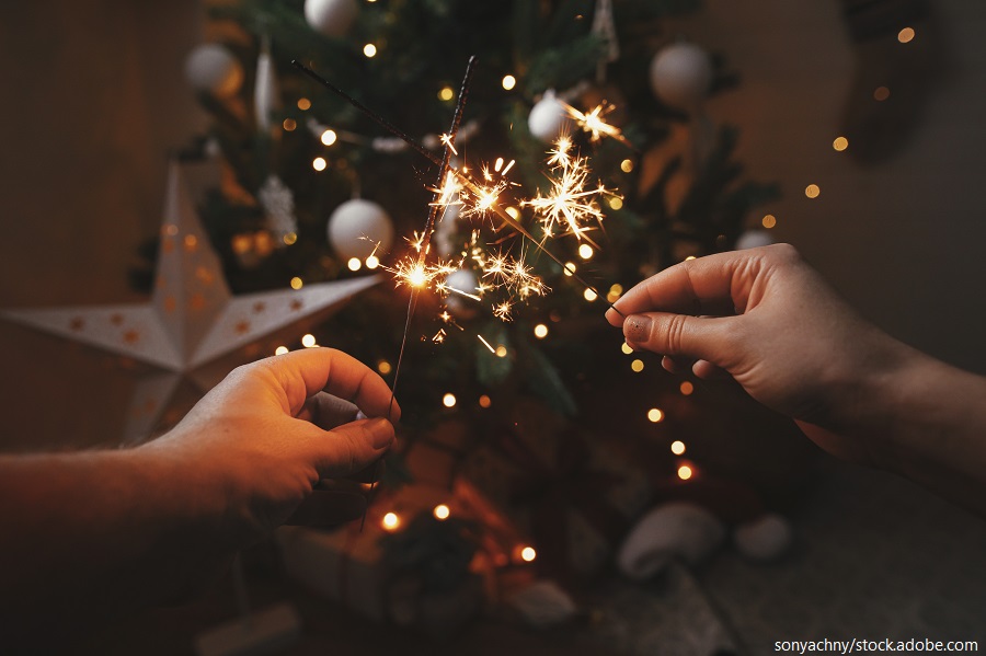 Zwei Hände halten Wunderkerzen vor einem Weihnachtsbaum, der mit Lichtern und Dekoration geschmückt ist.